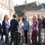 Eine Gruppe Jugendlicher und ein Erwachsener stehen auf einem Balkon, blicken in die Kamera und lächeln, während im Hintergrund historische Gebäude und ein Hügel zu sehen sind.