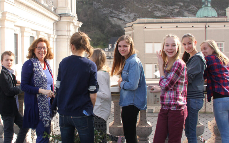 Eine Gruppe Jugendlicher und ein Erwachsener stehen auf einem Balkon, blicken in die Kamera und lächeln, während im Hintergrund historische Gebäude und ein Hügel zu sehen sind.