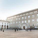 Ein großer, offener Platz mit Menschengruppen vor einem historischen, mehrstöckigen Gebäude mit bogenförmigen Eingängen und verzierten Fenstern.