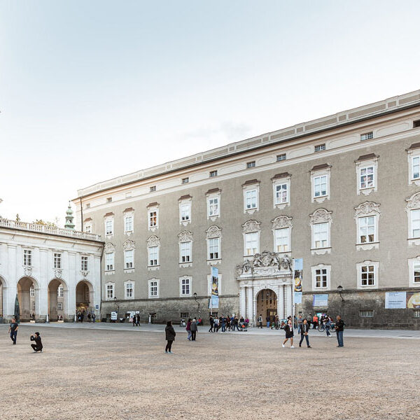 Ein großer, offener Platz mit Menschengruppen vor einem historischen, mehrstöckigen Gebäude mit bogenförmigen Eingängen und verzierten Fenstern.