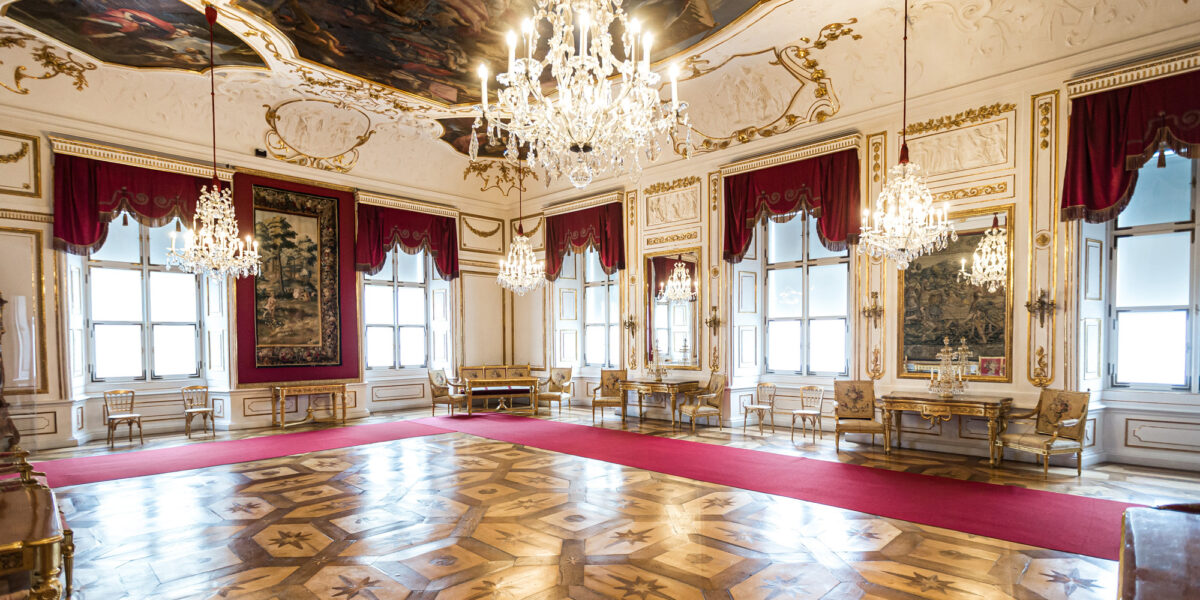 Opulenter Palastsaal mit kunstvollen Kronleuchtern, roten Vorhängen, Parkettböden, goldverzierten Wänden, großen Spiegeln und aufwändigen Deckengemälden.