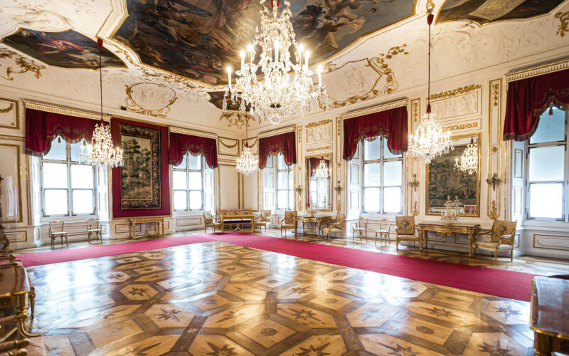 Opulenter Palastsaal mit kunstvollen Kronleuchtern, roten Vorhängen, Parkettböden, goldverzierten Wänden, großen Spiegeln und aufwändigen Deckengemälden.