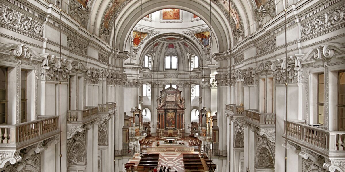 Barockes Kircheninnere mit verschnörkelten Bögen, Säulen, dekorativen Decken und einer Gruppe von Menschen, die sich vorne am Altar versammelt haben.