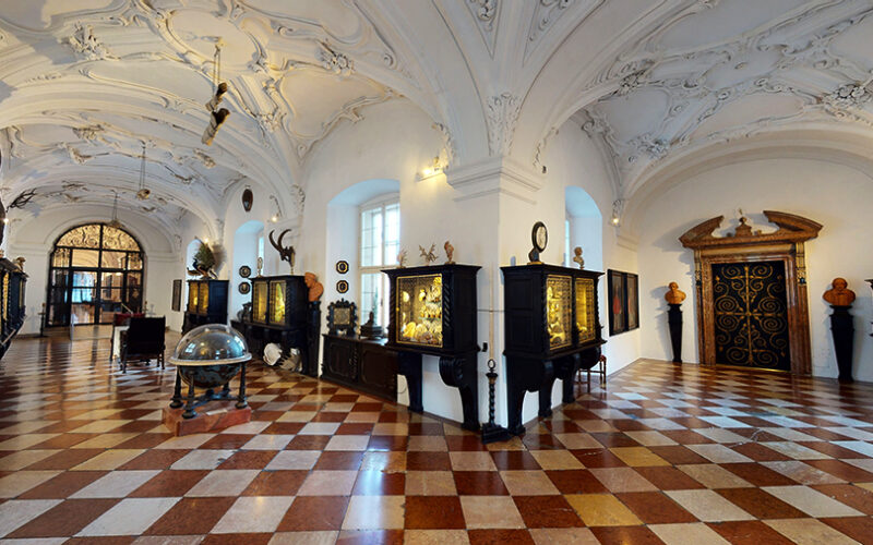 Verschnörkelter Museumssaal mit gewölbter, verzierter Decke, kariertem Boden, Vitrinen und antiken Globen und Artefakten an den Wänden.