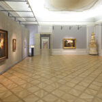 Geräumige Kunstgalerie mit Parkettboden, grauen Wänden und mehreren gerahmten Gemälden; in der hinteren Ecke befindet sich ein verschnörkelter Keramikofen.