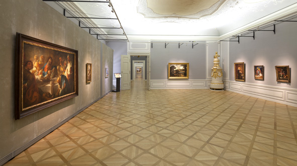 Geräumige Kunstgalerie mit Parkettboden, grauen Wänden und mehreren gerahmten Gemälden; in der hinteren Ecke befindet sich ein verschnörkelter Keramikofen.