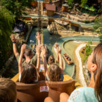 Eine Gruppe von Menschen mit erhobenen Händen fährt in einem Vergnügungspark eine Wasserrutsche steil hinunter, wobei das Wasser und die Parklandschaft unten sichtbar sind.