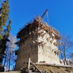 Ein im Bau befindlicher Steinturm, umgeben von Gerüsten und Holzstützen, mit Bäumen und einem klaren blauen Himmel im Hintergrund.