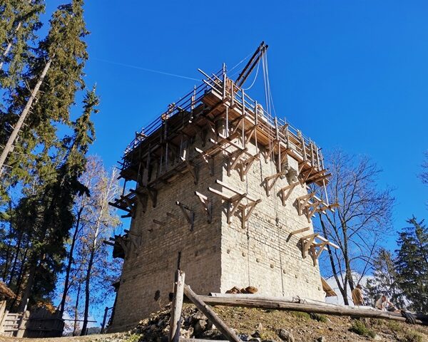 Ein im Bau befindlicher Steinturm, umgeben von Gerüsten und Holzstützen, mit Bäumen und einem klaren blauen Himmel im Hintergrund.