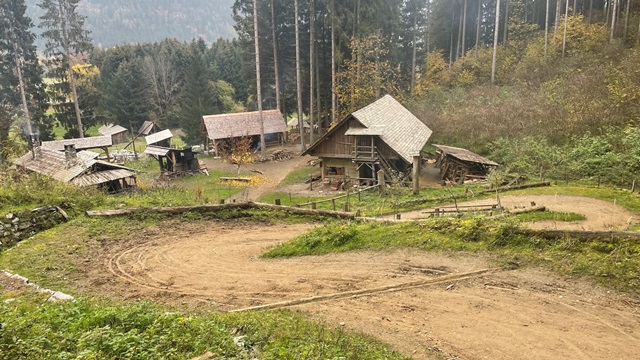 Eine Ansammlung von Holzhütten und Schuppen steht auf einem grasbewachsenen Hügel, umgeben von hohen Bäumen und einem Feldweg, der sich im Vordergrund schlängelt.
