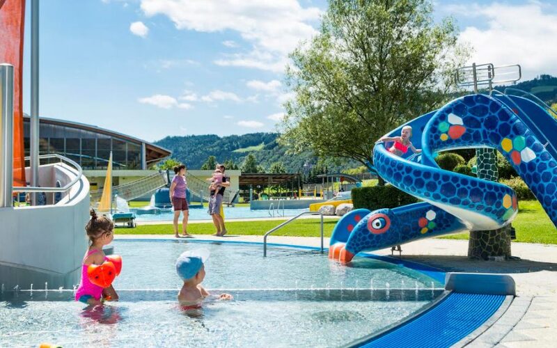 Kinder spielen in einem flachen Außenbecken neben einer blauen Schlangenrutsche, während Erwachsene an einem sonnigen Tag im Hintergrund zuschauen.