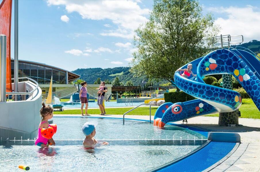Kinder spielen in einem flachen Außenbecken neben einer blauen Schlangenrutsche, während Erwachsene an einem sonnigen Tag im Hintergrund zuschauen.