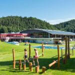 Kinder spielen auf einem hölzernen Hindernisparcours auf einer Wiese in der Nähe eines Freibads, mit einem großen modernen Gebäude und bewaldeten Hügeln im Hintergrund.
