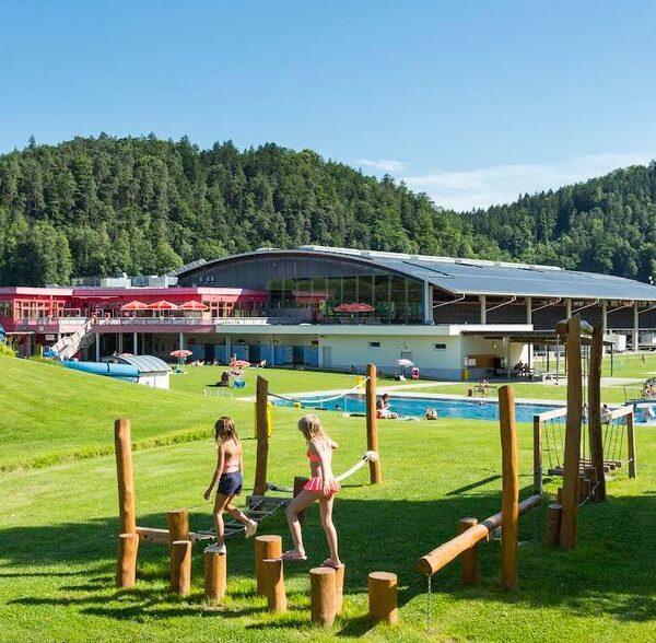 Kinder spielen auf einem hölzernen Hindernisparcours auf einer Wiese in der Nähe eines Freibads, mit einem großen modernen Gebäude und bewaldeten Hügeln im Hintergrund.