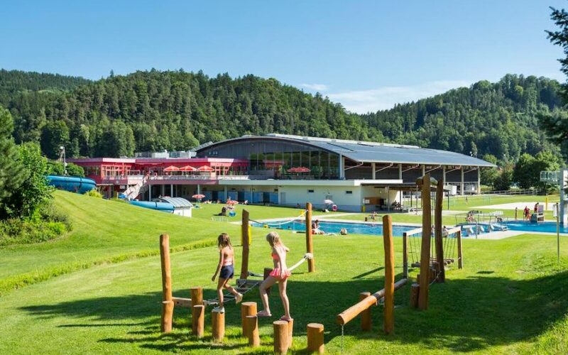Kinder spielen auf einem hölzernen Hindernisparcours auf einer Wiese in der Nähe eines Freibads, mit einem großen modernen Gebäude und bewaldeten Hügeln im Hintergrund.