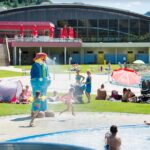 Familien und Kinder erfreuen sich an einem grasbewachsenen Außenbereich mit Pools, Zelten und einer großen Zeichentrickfiguren-Statue in der Nähe eines modernen Gebäudes mit großen Fenstern.