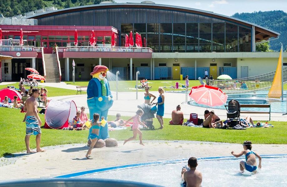 Familien und Kinder erfreuen sich an einem grasbewachsenen Außenbereich mit Pools, Zelten und einer großen Zeichentrickfiguren-Statue in der Nähe eines modernen Gebäudes mit großen Fenstern.
