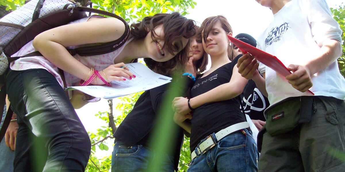 Eine Gruppe Teenager steht im Freien im Grünen, einige halten Papier und Stifte in den Händen und scheinen an einer Outdoor-Aktivität oder einem Studium teilzunehmen.
