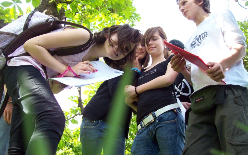 Eine Gruppe Teenager steht im Freien im Grünen, einige halten Papier und Stifte in den Händen und scheinen an einer Outdoor-Aktivität oder einem Studium teilzunehmen.