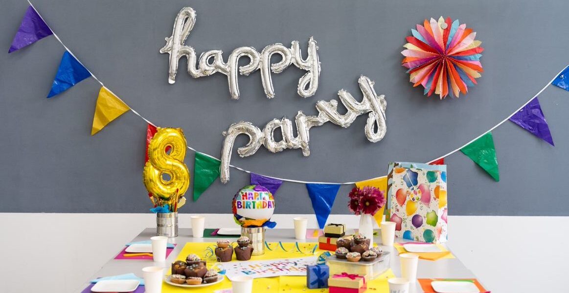 Ein geschmückter Geburtstagstisch mit bunten Tassen, Tellern, Luftballons, Muffins, einer Torte, Geschenken und einem "Happy Party"-Ballonbanner an der Wand.