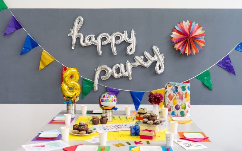 Ein geschmückter Geburtstagstisch mit bunten Tassen, Tellern, Luftballons, Muffins, einer Torte, Geschenken und einem "Happy Party"-Ballonbanner an der Wand.