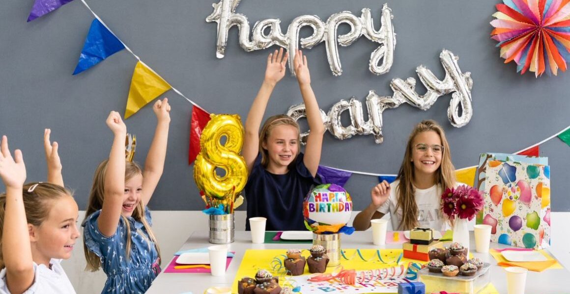 Vier Kinder sitzen an einem Tisch mit Geburtstagsdekoration, Muffins und Geschenken und heben feierlich ihre Arme unter einem "Happy Birthday"-Ballonbanner.