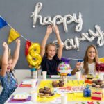 Vier Kinder sitzen an einem Tisch mit Geburtstagsdekoration, Muffins und Geschenken und heben feierlich ihre Arme unter einem "Happy Birthday"-Ballonbanner.