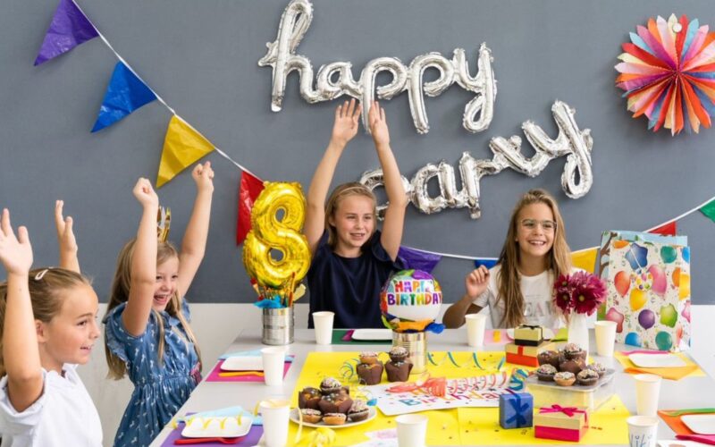 Vier Kinder sitzen an einem Tisch mit Geburtstagsdekoration, Muffins und Geschenken und heben feierlich ihre Arme unter einem "Happy Birthday"-Ballonbanner.