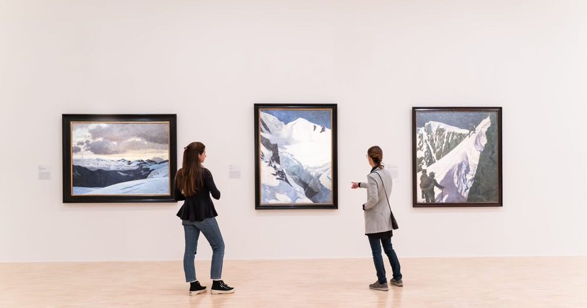 Zwei Personen stehen in einer Kunstgalerie und betrachten drei gerahmte Landschaftsgemälde von verschneiten Bergen an einer weißen Wand.