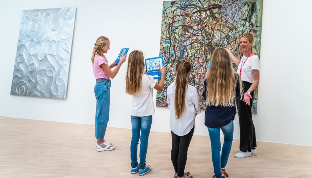 Vier Kinder mit Tablets stehen neben einer Frau vor abstrakten Bildern in einer Kunstgalerie mit hellen Böden und Wänden.