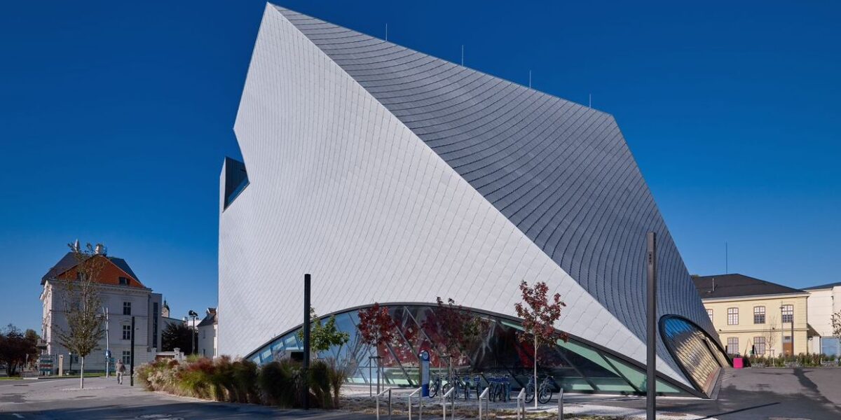 Ein modernes Gebäude mit kantiger, geometrischer Architektur, einem silbernen, kachelähnlichen Äußeren und großen geschwungenen Glasfenstern steht in einem städtischen Gebiet unter einem klaren blauen Himmel.