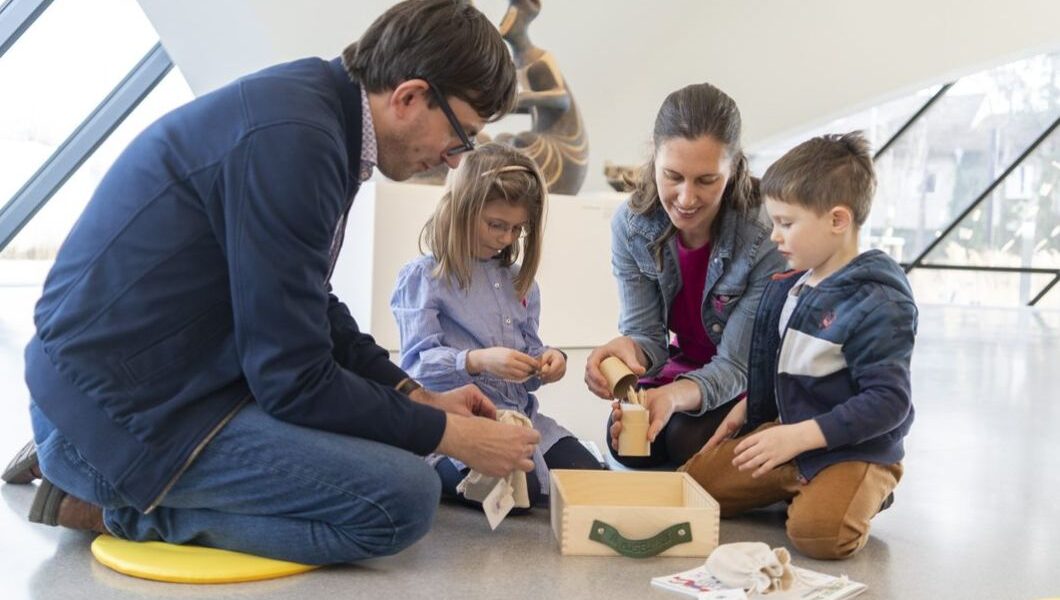 Zwei Erwachsene und zwei Kinder sitzen in einem hellen, modernen Raum mit großen Fenstern auf dem Boden und bauen Holzobjekte aus einer Kiste zusammen.