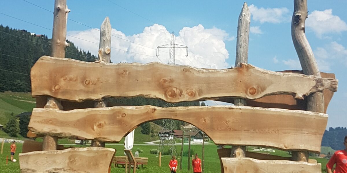 Große Holzkonstruktion mit unregelmäßig horizontal gestapelten Brettern und einem zentralen Bogen auf einer Wiese mit Menschen und Hügeln im Hintergrund.