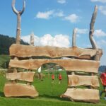 Große Holzkonstruktion mit unregelmäßig horizontal gestapelten Brettern und einem zentralen Bogen auf einer Wiese mit Menschen und Hügeln im Hintergrund.