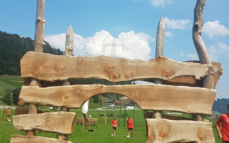 Große Holzkonstruktion mit unregelmäßig horizontal gestapelten Brettern und einem zentralen Bogen auf einer Wiese mit Menschen und Hügeln im Hintergrund.