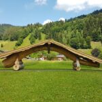Auf einer Wiese steht eine Holzkonstruktion mit schrägem Dach, im Hintergrund sind grüne Hügel, Bäume und verstreute Häuser unter einem blauen Himmel zu sehen.