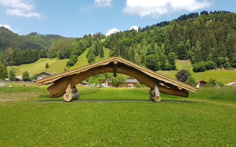 Auf einer Wiese steht eine Holzkonstruktion mit schrägem Dach, im Hintergrund sind grüne Hügel, Bäume und verstreute Häuser unter einem blauen Himmel zu sehen.