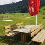 Hölzerner Picknicktisch mit Bänken und einem geschlossenen roten Sonnenschirm auf einer Wiese, mit Hügeln und Bäumen im Hintergrund unter einem teilweise bewölkten Himmel.