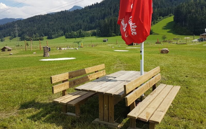Hölzerner Picknicktisch mit Bänken und einem geschlossenen roten Sonnenschirm auf einer Wiese, mit Hügeln und Bäumen im Hintergrund unter einem teilweise bewölkten Himmel.