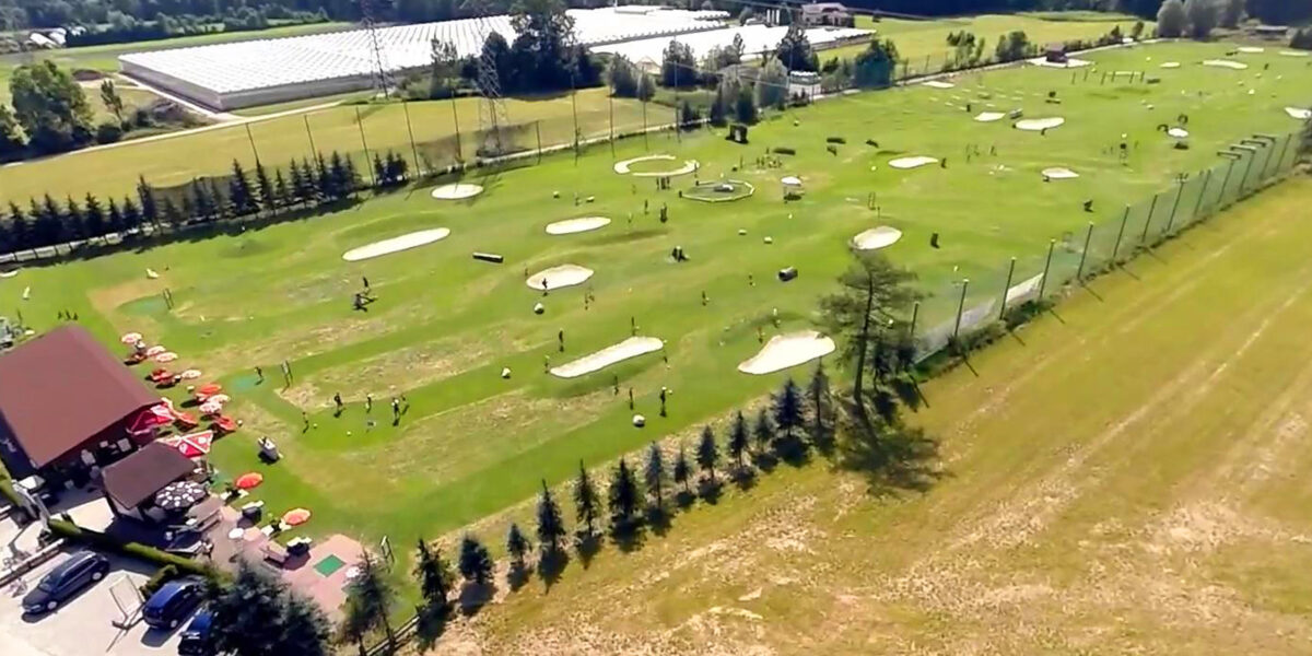 Luftaufnahme eines großen Golfplatzes mit mehreren Übungsgrüns, Sandbunkern und Zielscheiben; an einem Rand sind Gebäude und geparkte Autos zu sehen.