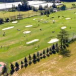 Luftaufnahme eines großen Golfplatzes mit mehreren Übungsgrüns, Sandbunkern und Zielscheiben; an einem Rand sind Gebäude und geparkte Autos zu sehen.