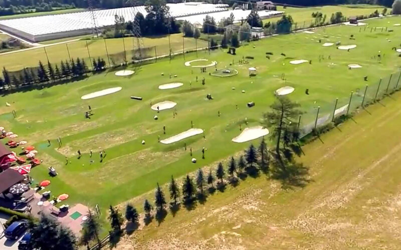 Luftaufnahme eines großen Golfplatzes mit mehreren Übungsgrüns, Sandbunkern und Zielscheiben; an einem Rand sind Gebäude und geparkte Autos zu sehen.
