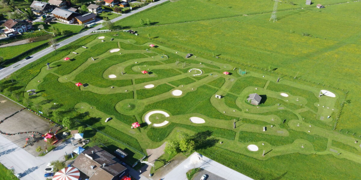 Luftaufnahme eines Minigolfplatzes mit gewundenen Wegen, kreisförmigen Grüns, Hindernissen und roten Sonnenschirmen, umgeben von Grasfeldern und nahe gelegenen Gebäuden.