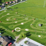 Luftaufnahme eines Minigolfplatzes mit gewundenen Wegen, kreisförmigen Grüns, Hindernissen und roten Sonnenschirmen, umgeben von Grasfeldern und nahe gelegenen Gebäuden.