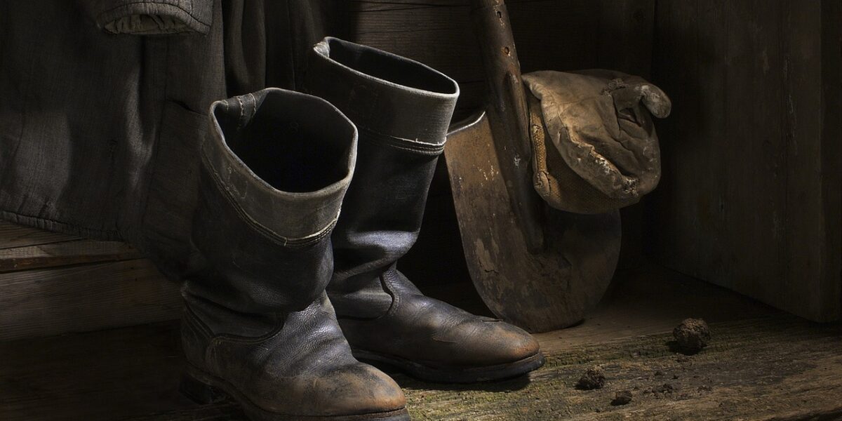 Ein Paar abgetragener schwarzer Stiefel, eine Schaufel mit Schlamm auf dem Blatt und ein Mantel, der an einer Holzwand hängt, mit Schmutzklumpen auf dem Boden.