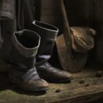 Ein Paar abgetragener schwarzer Stiefel, eine Schaufel mit Schlamm auf dem Blatt und ein Mantel, der an einer Holzwand hängt, mit Schmutzklumpen auf dem Boden.