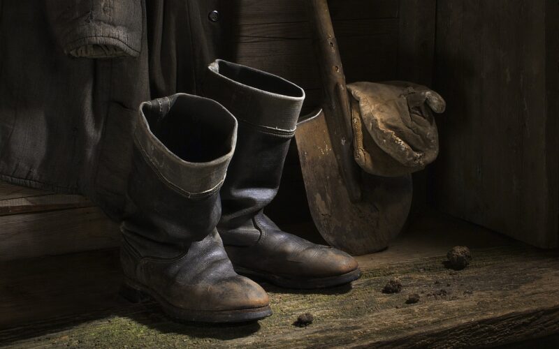 Ein Paar abgetragener schwarzer Stiefel, eine Schaufel mit Schlamm auf dem Blatt und ein Mantel, der an einer Holzwand hängt, mit Schmutzklumpen auf dem Boden.