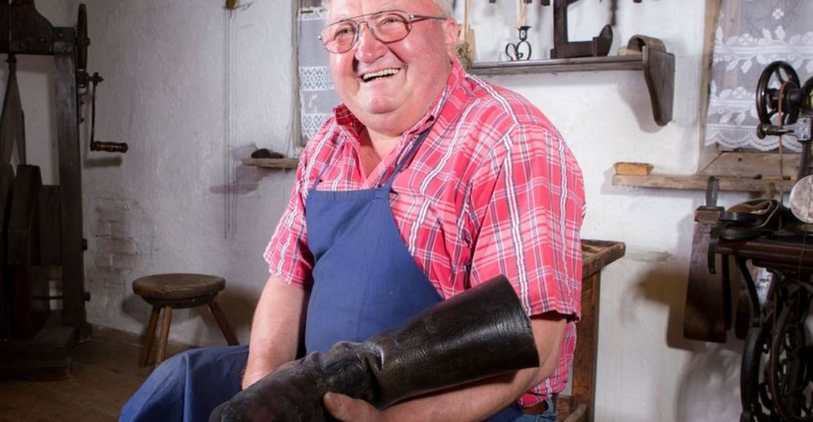 Ein älterer Mann mit Brille und blauer Schürze hält einen großen schwarzen Stiefel in der Hand. Er sitzt in einer Werkstatt, hinter ihm hängen Werkzeuge an der Wand.