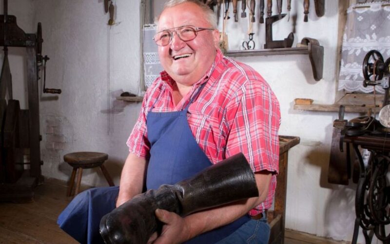 Ein älterer Mann mit Brille und blauer Schürze hält einen großen schwarzen Stiefel in der Hand. Er sitzt in einer Werkstatt, hinter ihm hängen Werkzeuge an der Wand.
