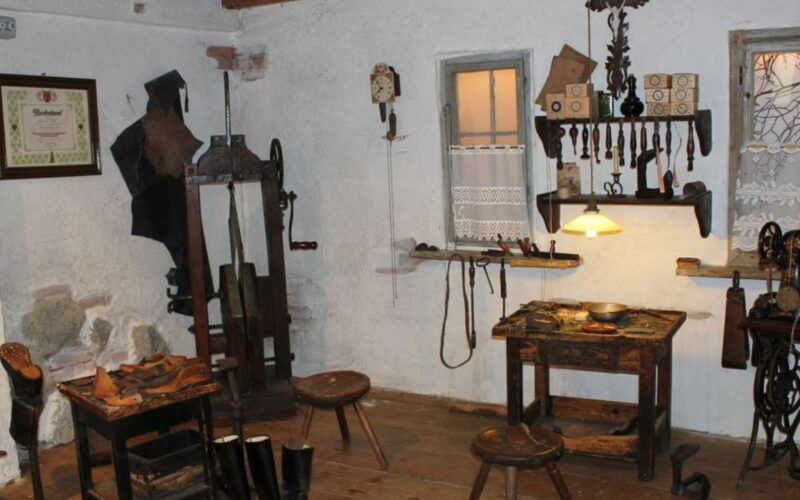 Eine alte Schusterwerkstatt mit Holzhockern, Werkzeugen an den Wänden, einer Nähmaschine, Schuhen und Werkbänken, beleuchtet von einer Hängelampe.